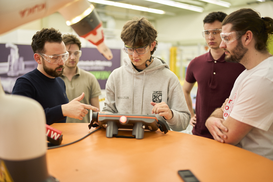 A trainer explains the use of a control device to students gathered around an industrial robot in the lab.