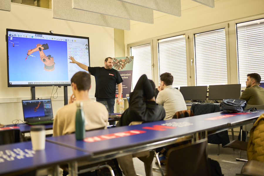 An instructor presents a robotic arm model on a large screen to students during a classroom session on industrial robotics.