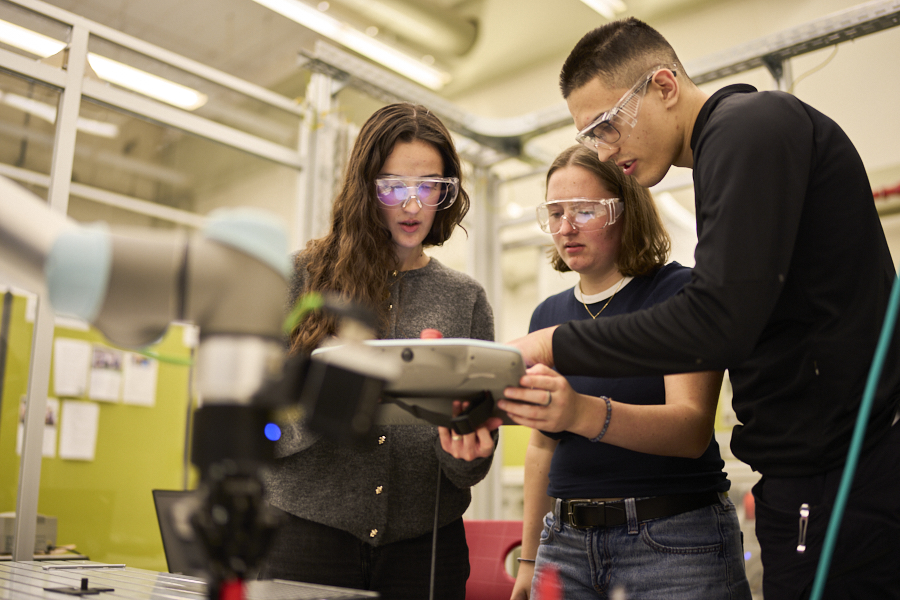 Three students work together with a control device next to industrial robotics equipment during a lab session.
