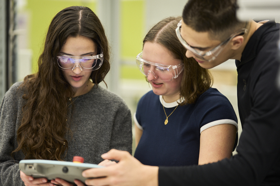 Three students in safety goggles look at a control device together during hands-on training in the robotics lab.