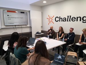 Participants seated around a table during a WP4 meeting in Lyon, listening and taking notes while a presentation slide is displayed on a screen.