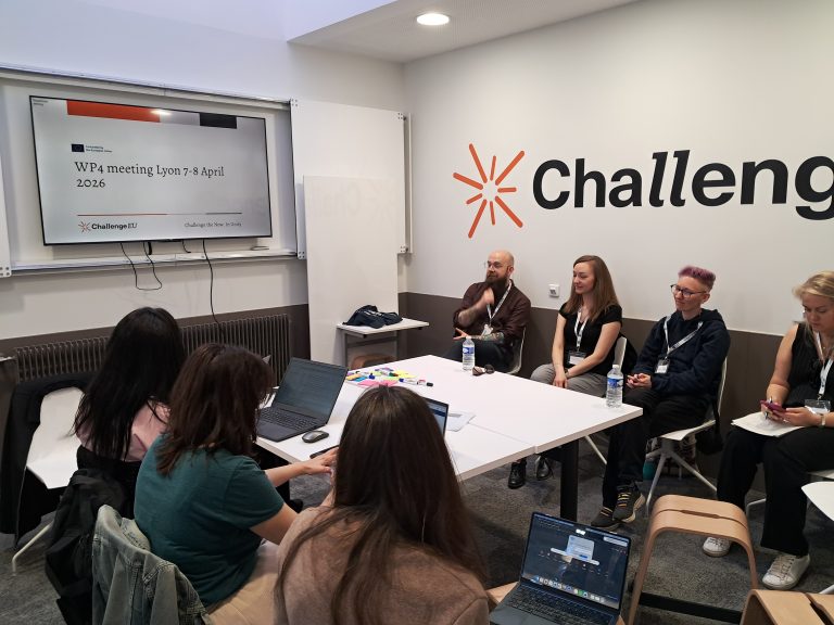 Participants seated around a table during a WP4 meeting in Lyon, listening and taking notes while a presentation slide is displayed on a screen.