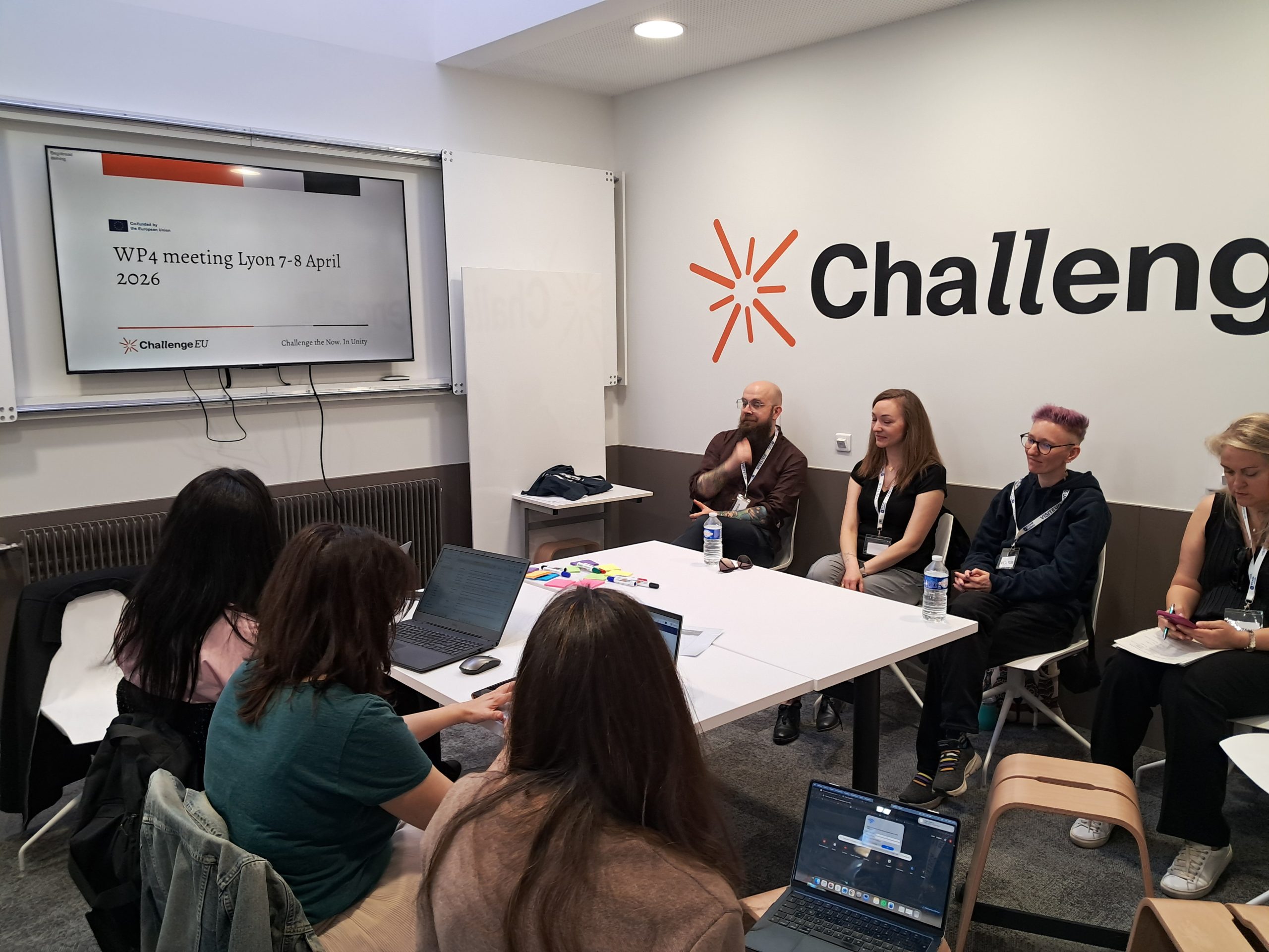 Participants seated around a table during a WP4 meeting in Lyon, listening and taking notes while a presentation slide is displayed on a screen.