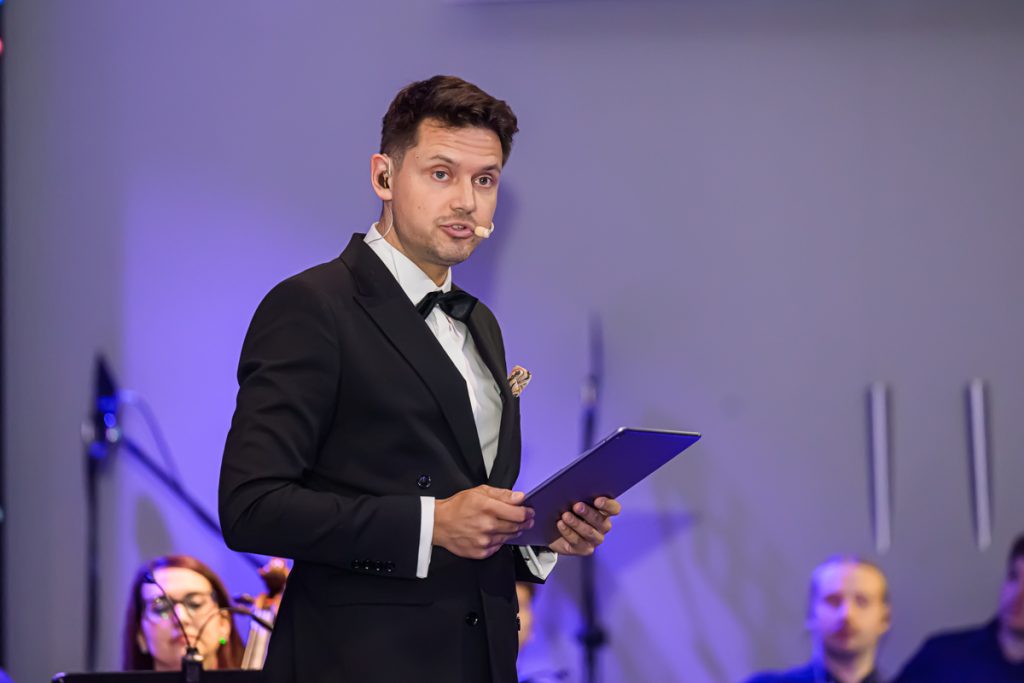 The host of the concert speaks on stage with the orchestra seated behind him.