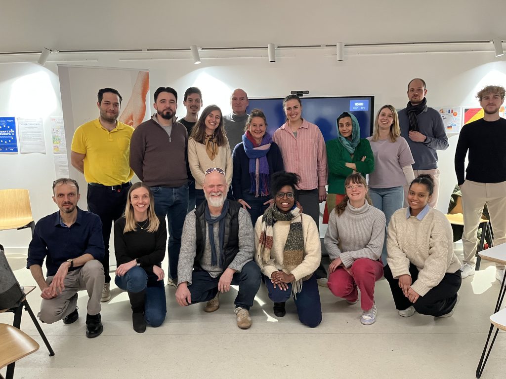Participants standing at the front of a workshop room, presenting ideas during a collaborative session, while others are seated and listening; a whiteboard with notes is visible in the background.