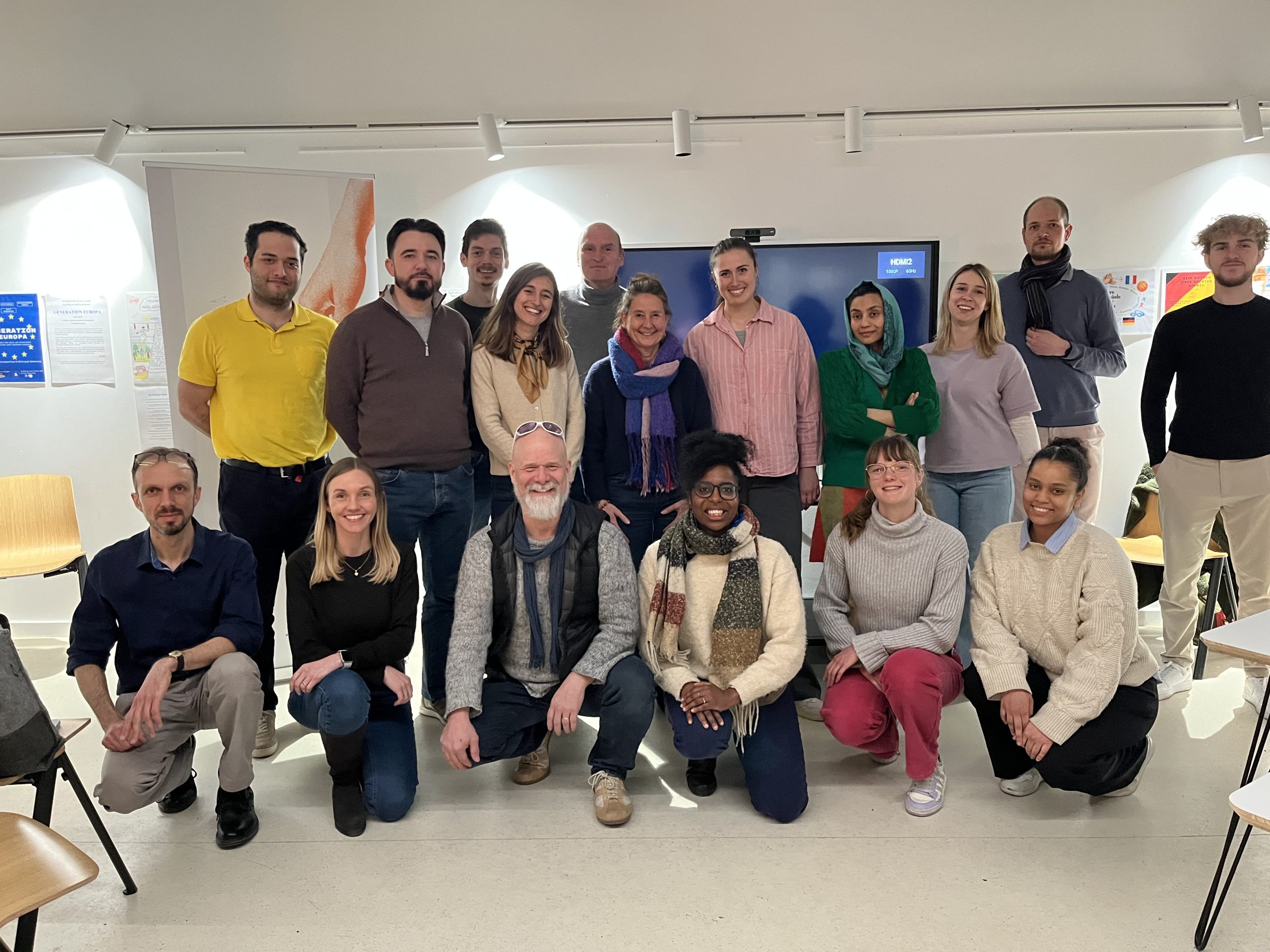 Participants standing at the front of a workshop room, presenting ideas during a collaborative session, while others are seated and listening; a whiteboard with notes is visible in the background.