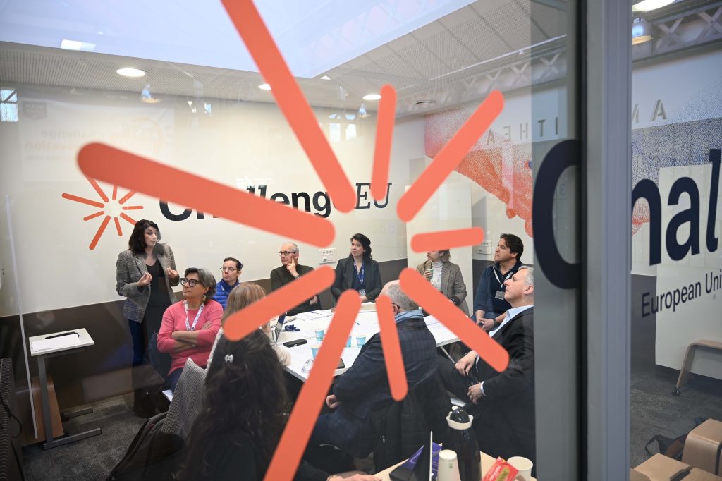 View through a glass wall showing a ChallengeEU meeting in progress, with a speaker addressing participants and the alliance’s visual identity visible in the foreground.