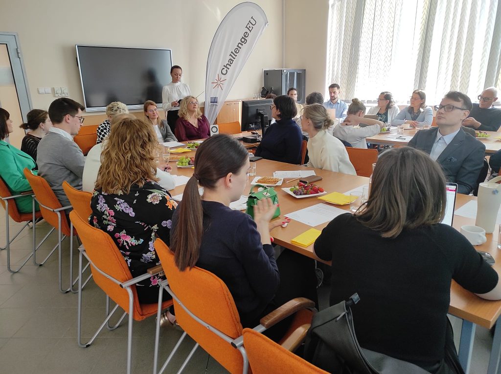 A group of participants seated around a conference table during a meeting at the University of Warmia and Mazury, listening to a speaker standing near a screen and a ChallengeEU banner.