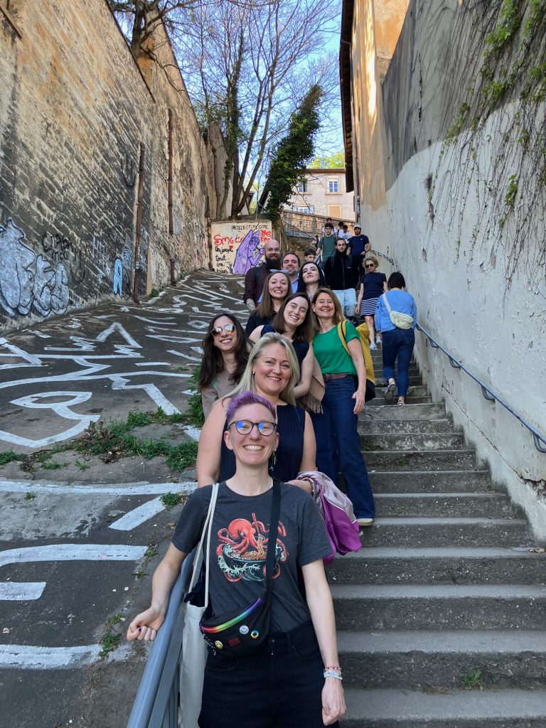 A group of ChallengeEU participants standing on outdoor stairs between old buildings with graffiti, smiling during a group photo in Lyon.
