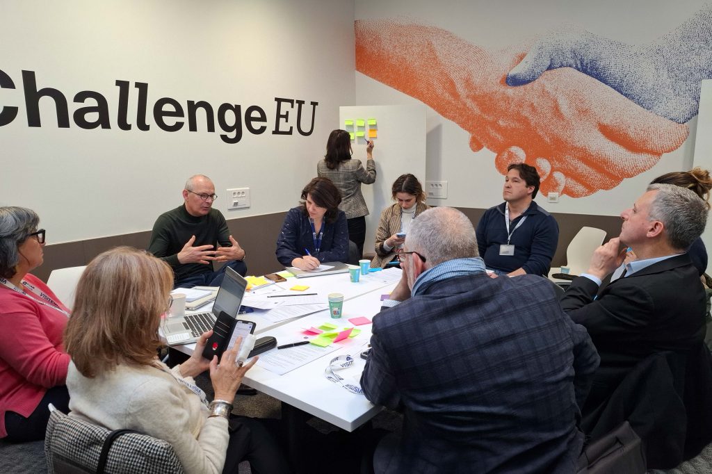Participants collaborating during a ChallengeEU workshop, taking notes and discussing ideas, with a visual of a handshake symbolising cooperation in the background.