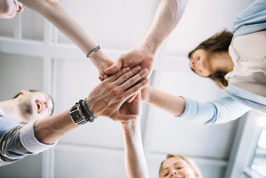 Group of people standing in a circle with their hands stacked together in the centre, symbolising teamwork, unity, and collaboration.
