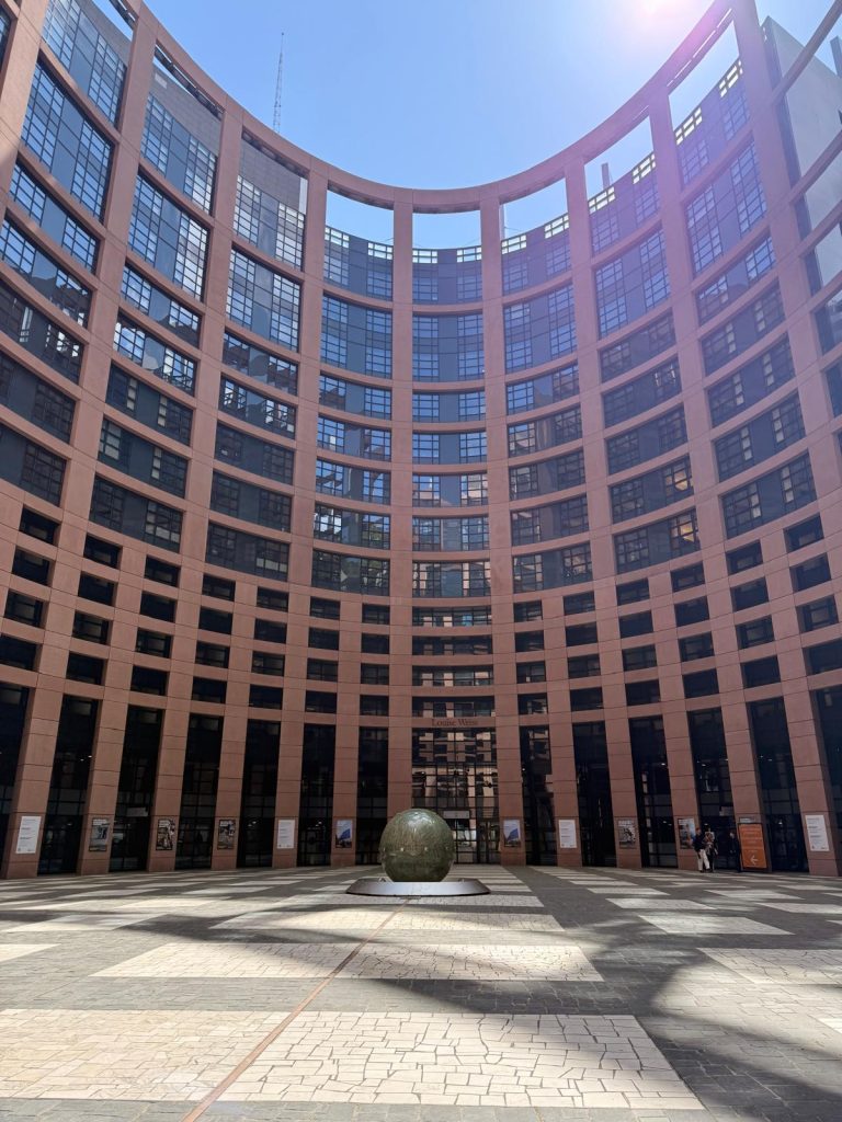 Exterior view of the Louise Weiss building of the European Parliament in Strasbourg, with its curved glass façade, sunlit courtyard and a spherical sculpture in the centre.