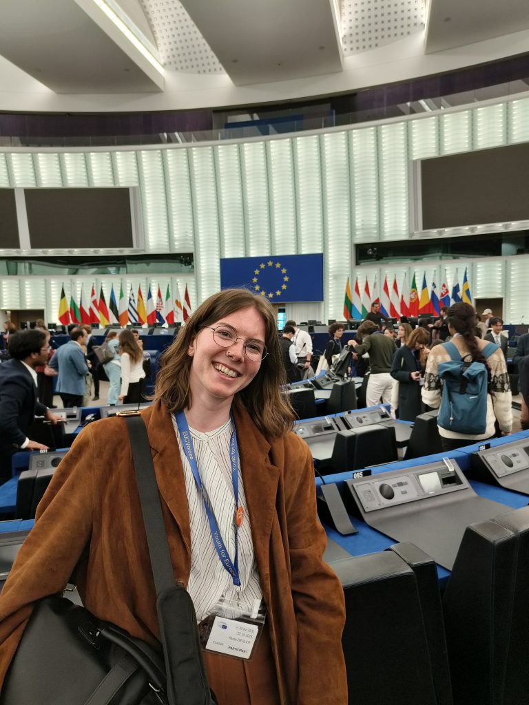 A smiling European Student Assembly 2026 participant stands inside the European Parliament hemicycle in Strasbourg, with delegates and EU flags visible in the background.