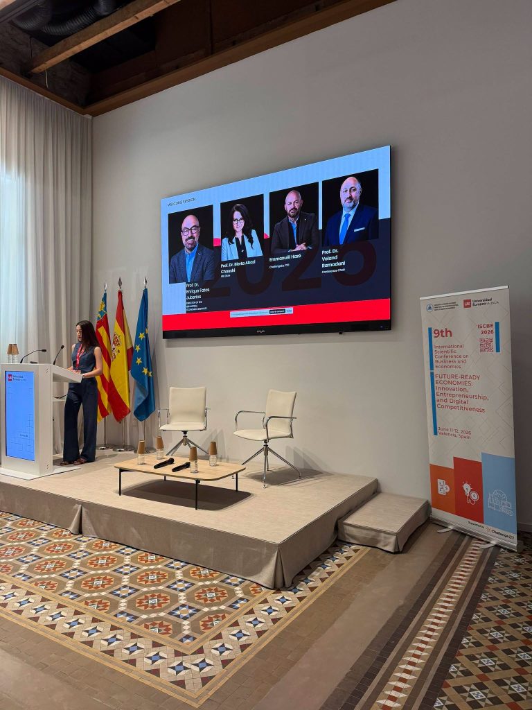 A speaker stands at a podium during the ISCBE 2026 Welcome Session, with a large screen showing the session speakers and an event banner nearby.