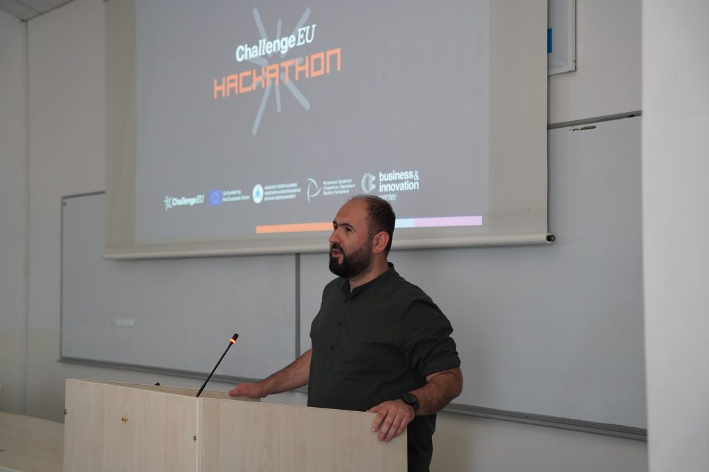 A speaker addresses the audience from a lectern in front of a screen displaying the ChallengeEU Hackathon logo.