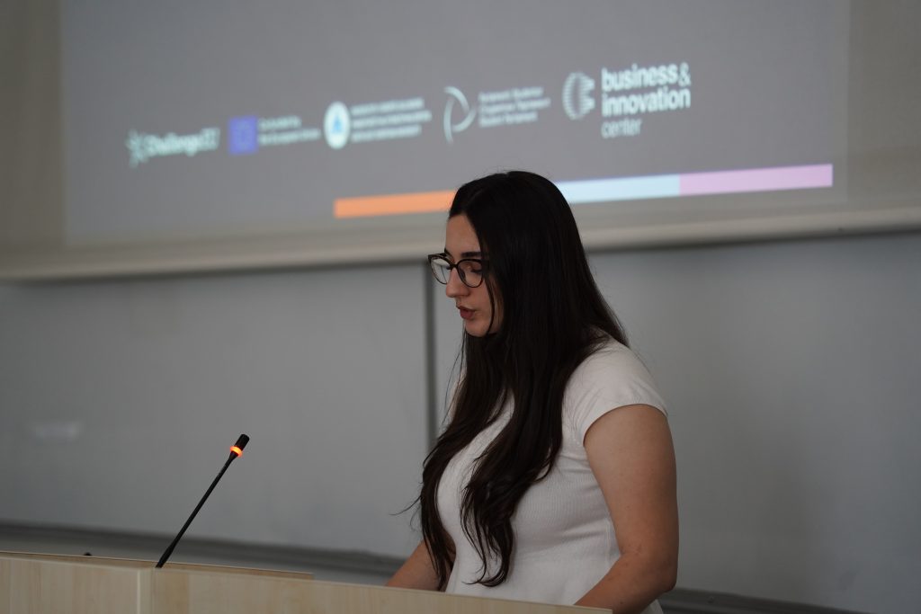 A hackathon participant presents a project at the lectern in front of a screen displaying the event logo.