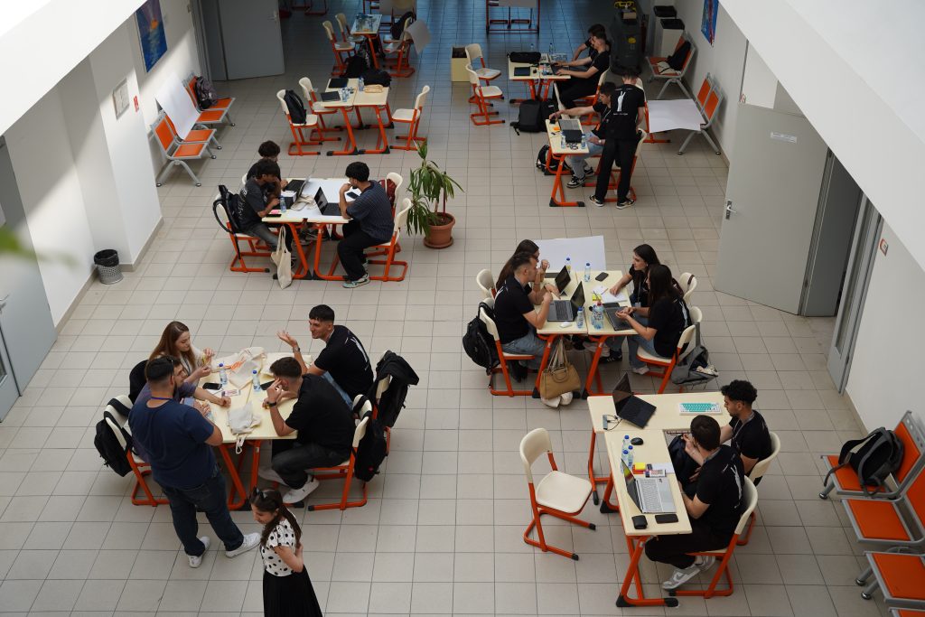 A top-down view of hackathon participants working in teams with laptops and notes in a spacious university hall.