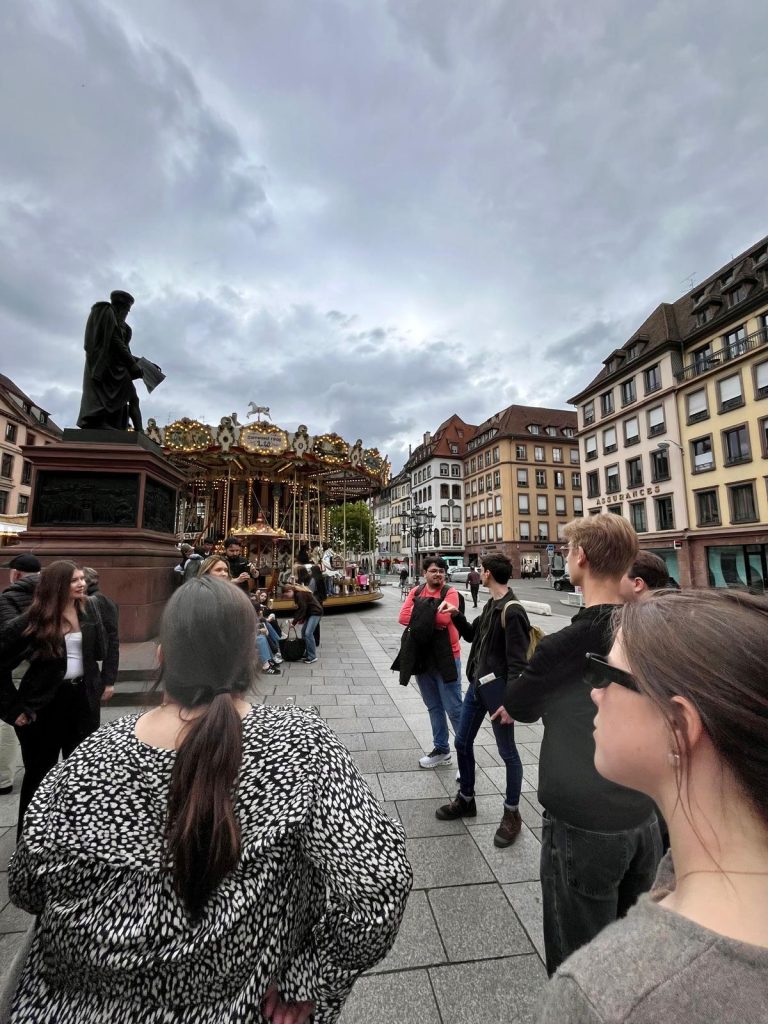 Students gather in a city square during a guided tour, with a monument and carousel nearby.
