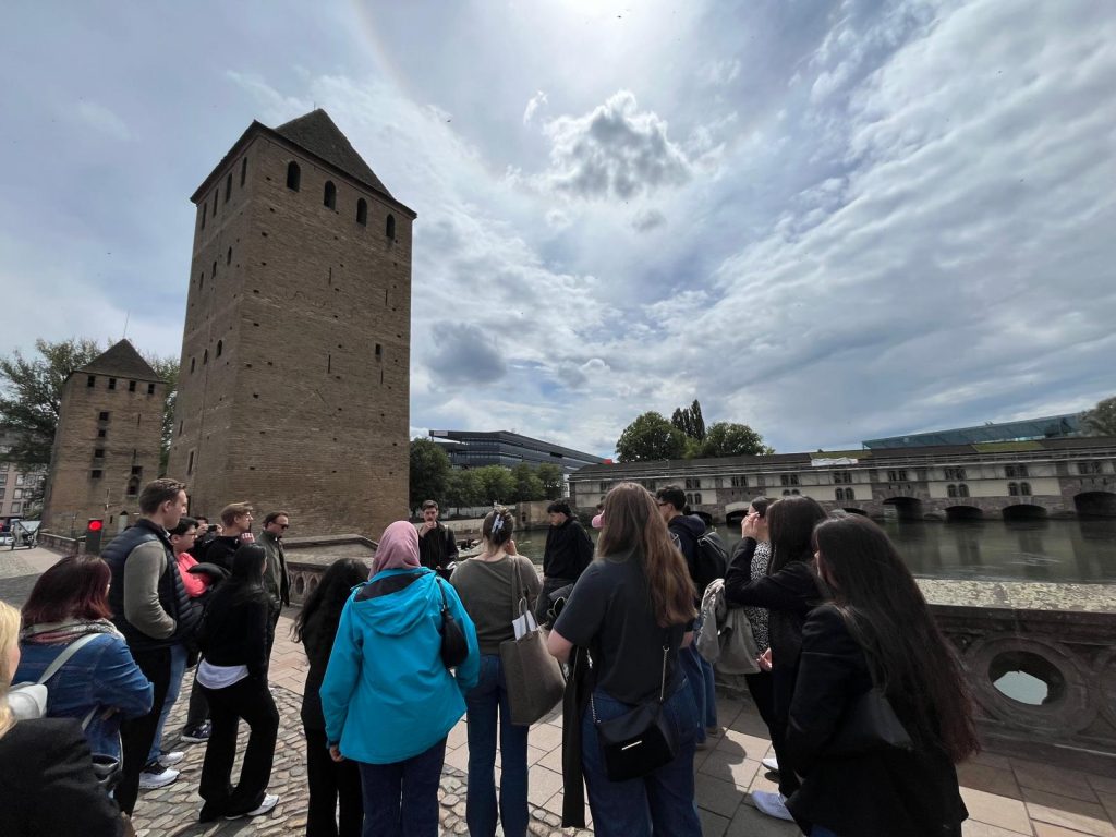 Students take part in a guided walk by the river, with historic towers and a bridge in the background.
