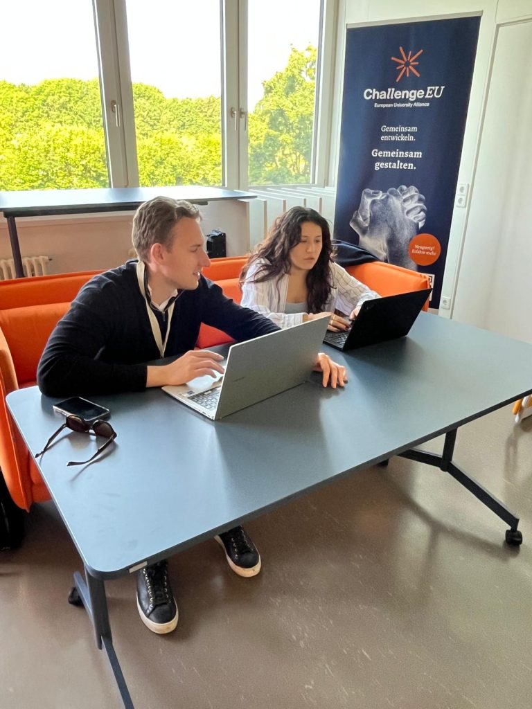 Two students work on laptops in front of a ChallengeEU banner.