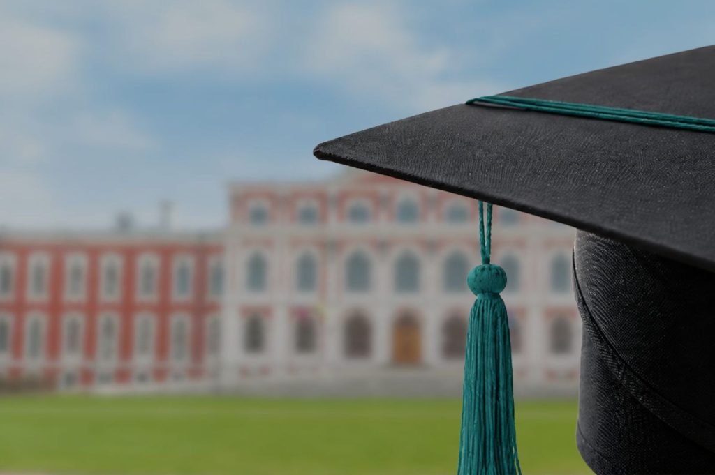 A close-up of a black graduation cap with a turquoise tassel, with a blurred university building in the background.