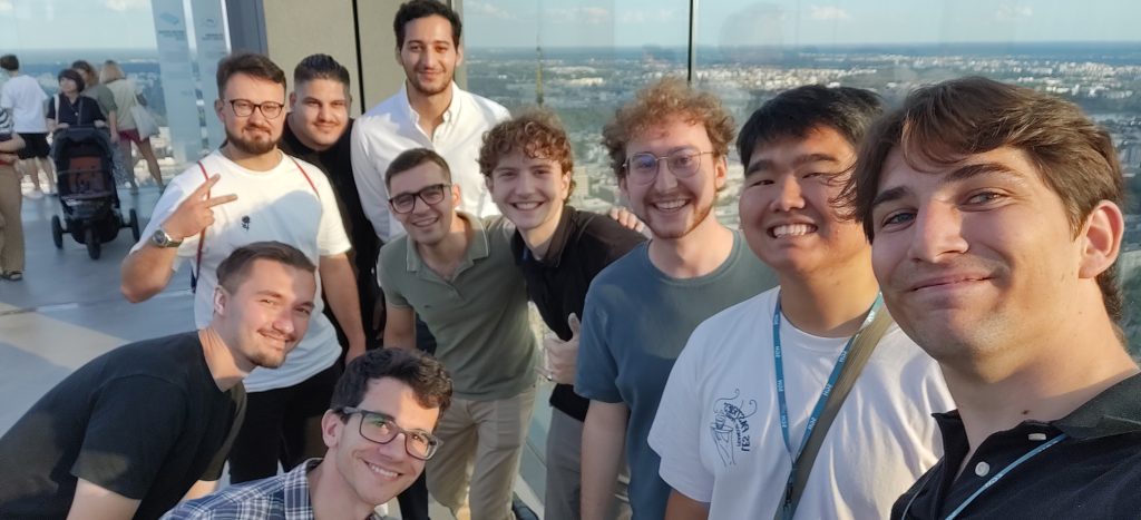 A group of student participants takes a selfie on a high observation deck overlooking Warsaw.