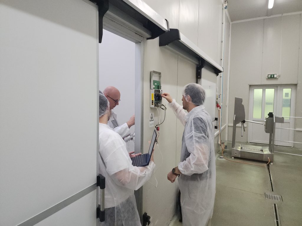 Three people wearing protective clothing and hairnets carry out sensor testing in a refrigeration facility. One person adjusts equipment mounted on the wall while another uses a laptop to monitor the process.